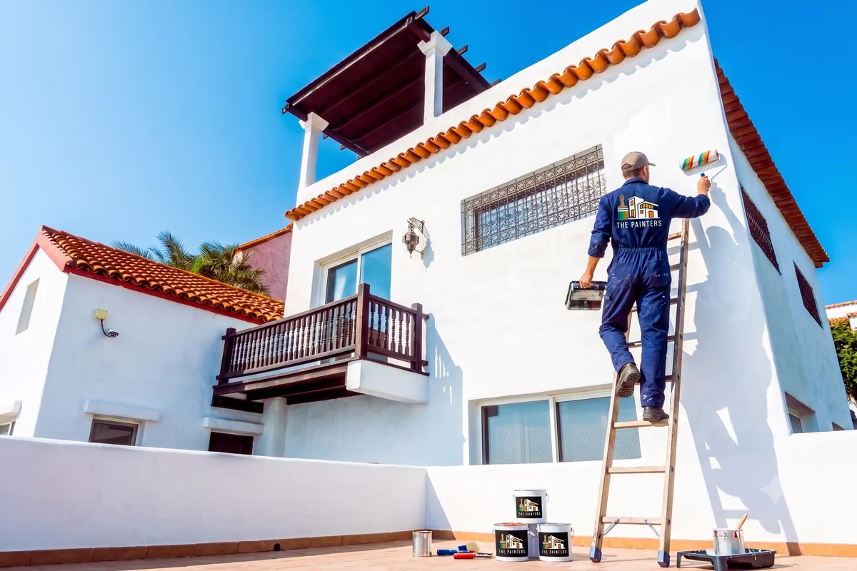 Pintor de ThePainters aplicando pintura en una fachada blanca, con botes de pintura profesional de primera calidad